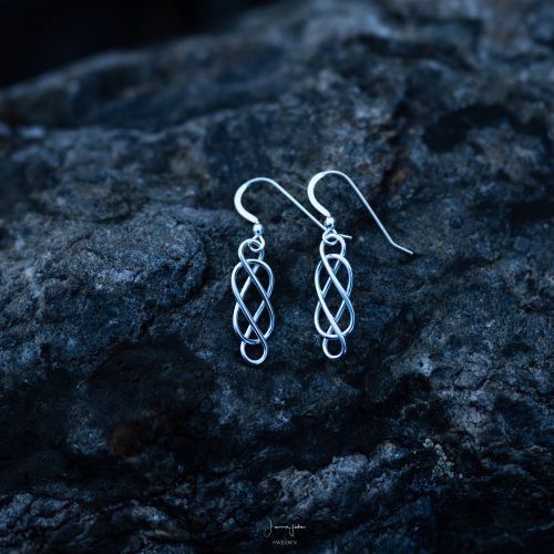 A pair of silver earrings with a celtic love knot laying on a dark rock