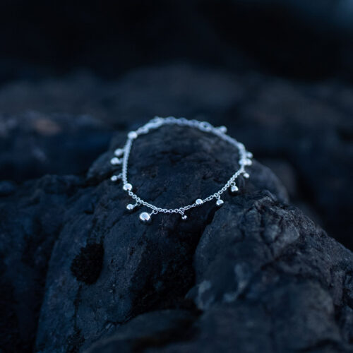 A sterling silver bead bracelet with polished silver beads in varying sizes on a delicate 925 sterling silver chain is laying on a dark rock